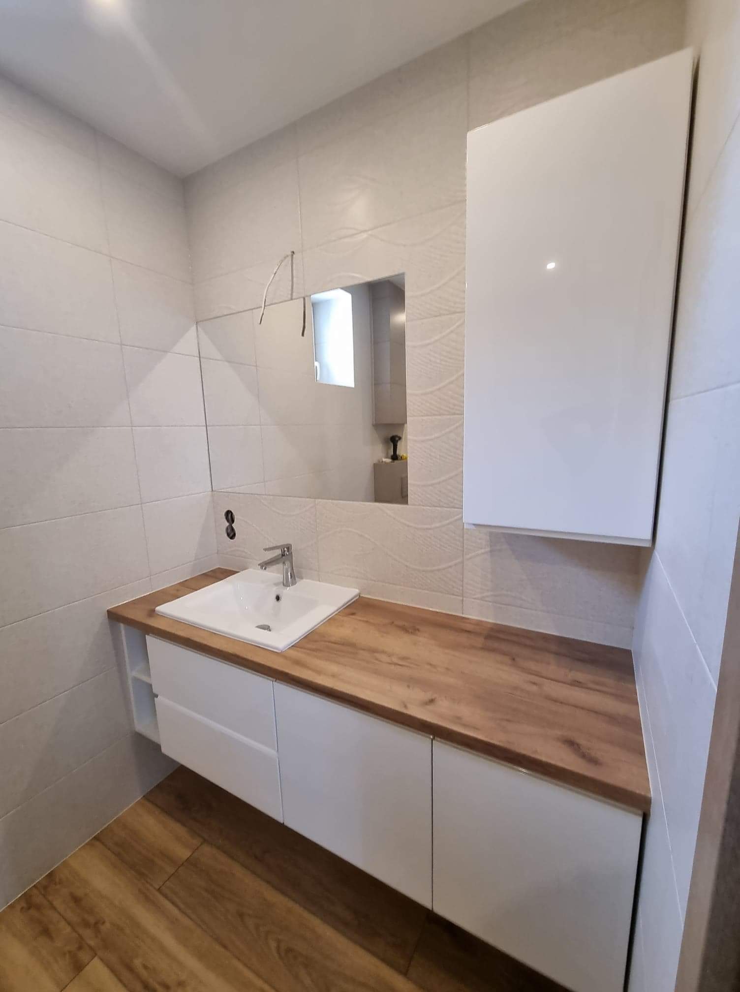 Nowoczesna łazienka w Kłocku z białymi szafkami pod umywalką z drewnianym blatem i wiszącą szafką nad umywalką, widok na lustro i wzorzyste płytki na ścianie.
