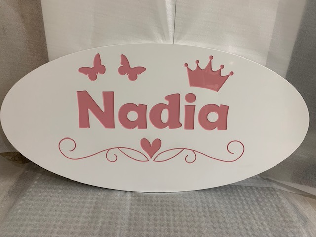Owalna tabliczka z białego drewna z imieniem 'Nadia' w kolorze różowym, ozdobiona motywami korony, motyli i serca z ornamentami.