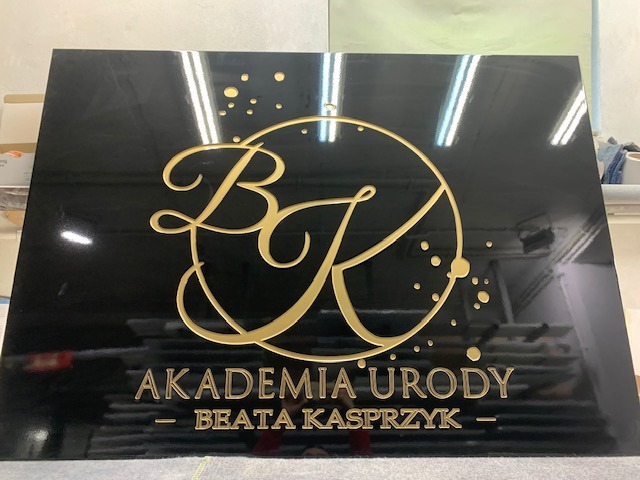 Czarna tablica z wyfrezowanym złotym logo 'BK Akademia Urody Beata Kasprzyk' w nowoczesnym stylu, widoczne refleksy świetlne na błyszczącej powierzchni.