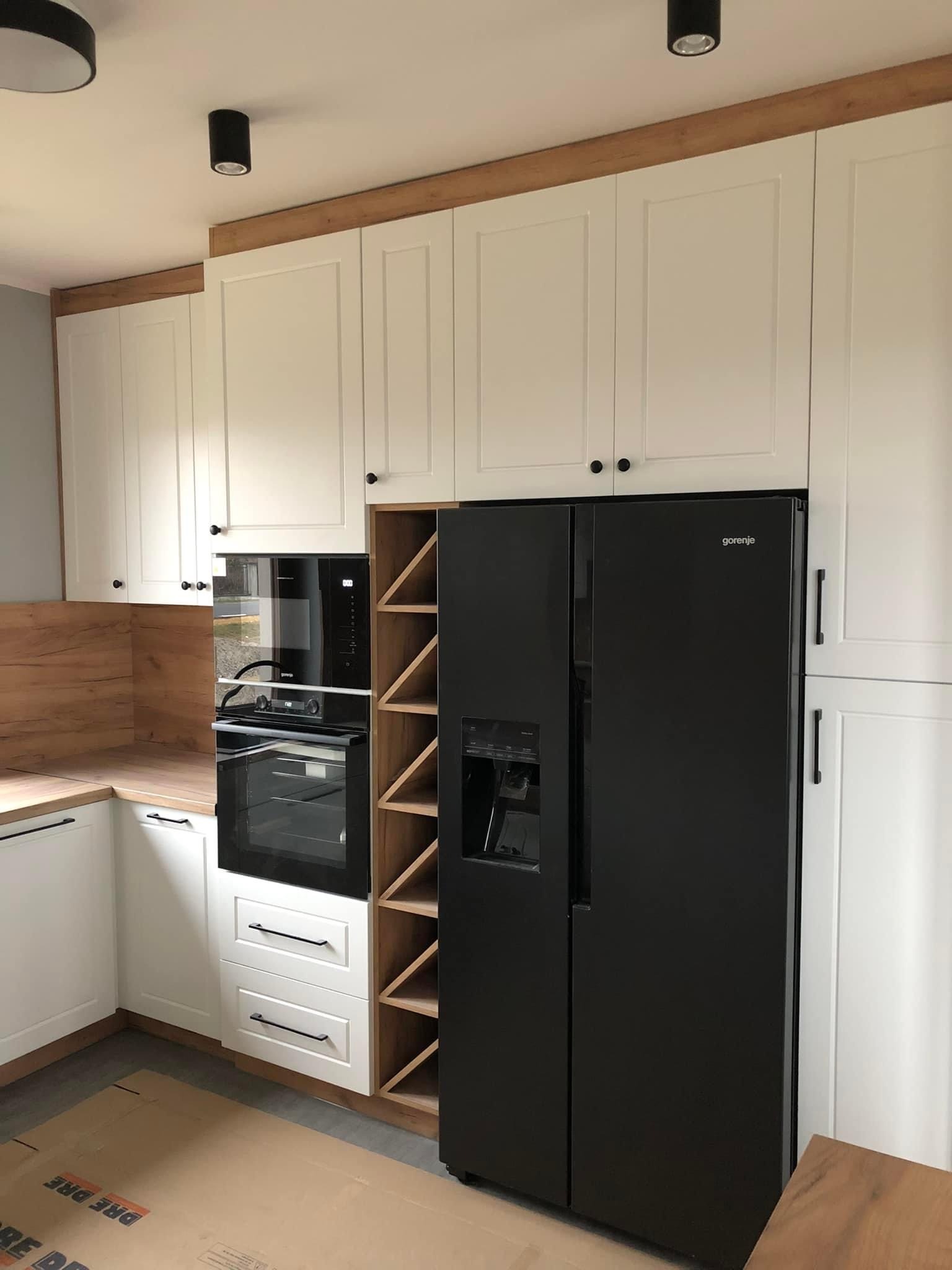 Nowoczesna, biała kuchnia z drewnianymi akcentami, czarnym sprzętem AGD i regałem na wino, oświetlona punktowymi lampami sufitowymi. Na podłodze widoczne kartony zabezpieczające.