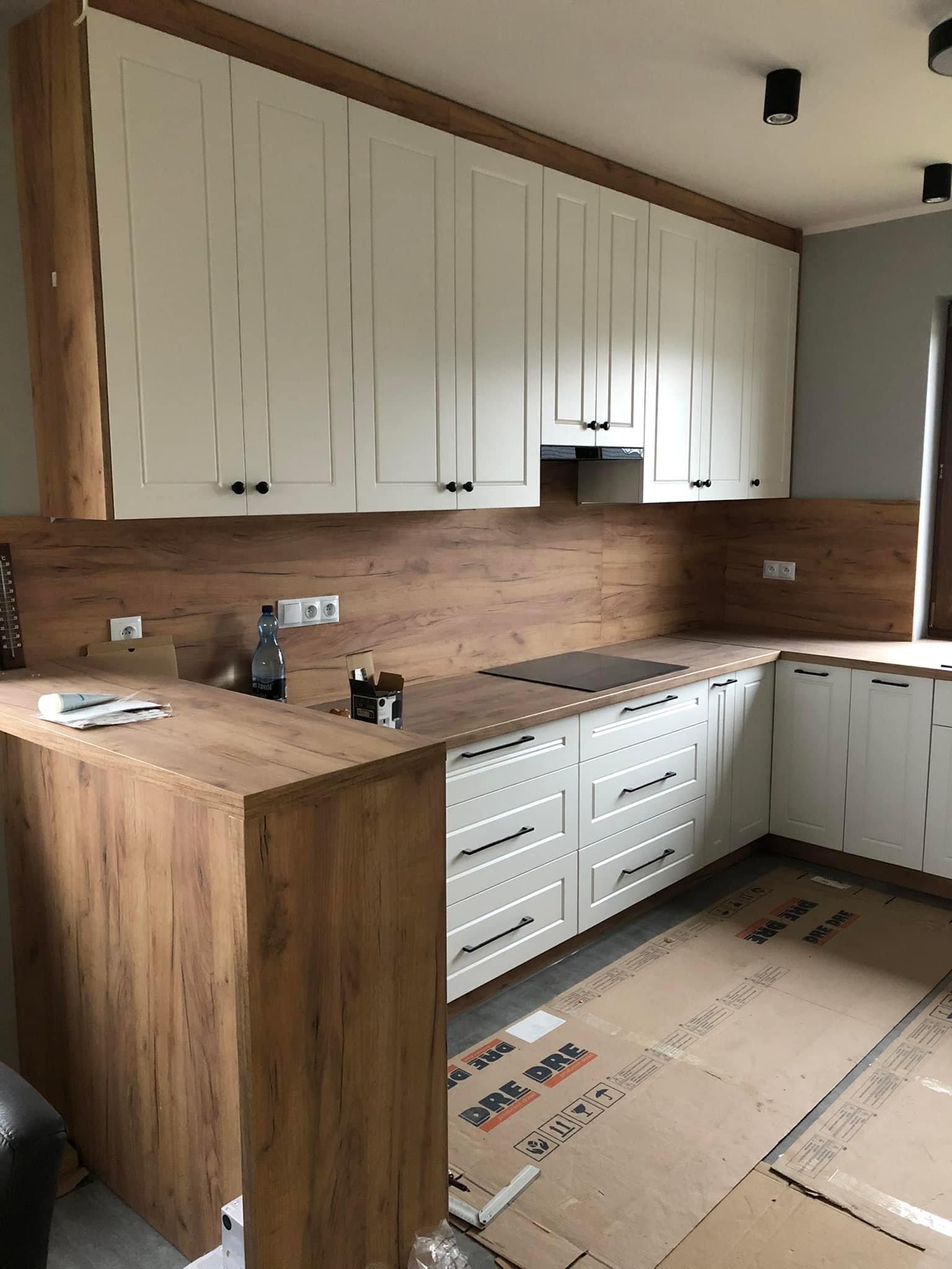 Nowoczesna kuchnia z białymi szafkami i drewnopodobnym blatem, widoczne kartony zabezpieczające podłogę podczas montażu, butelka wody i akcesoria na blacie.