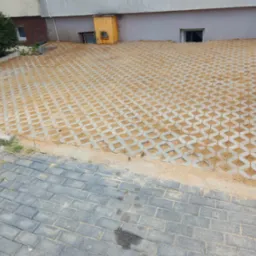 Ułożona nawierzchnia z betonowych ażurowych płyt wypełnionych piaskiem, przechodząca w tradycyjną kostkę brukową, widoczny fragment budynku z żółtą skrzynką gazową.