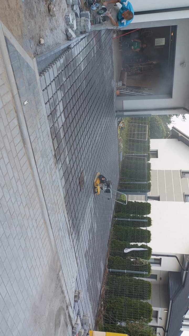 Układanie kostki brukowej na podjeździe, widoczna zagęszczarka wibrująca, w tle mężczyzna tnący kostkę szlifierką kątową w garażu.