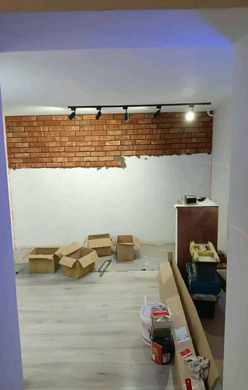 Ściana w trakcie wykańczania z częściowo położoną okładziną z cegły klinkierowej, widoczne kartony, narzędzia i listwy przypodłogowe na podłodze z paneli.