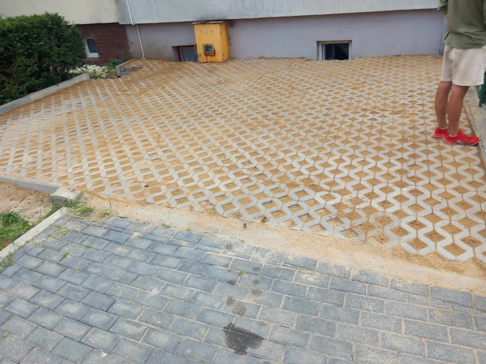 Ułożona nawierzchnia z betonowych ażurowych płyt wypełnionych piaskiem, przechodząca w tradycyjną kostkę brukową, widoczny fragment budynku z żółtą skrzynką gazową.