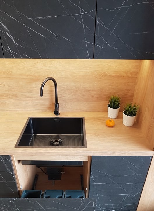 Nowoczesna kuchnia z czarnym zlewem, baterią i blatem z jasnego drewna. Otwarta szafka ukazuje system segregacji odpadów. Na blacie dekoracje: dwie doniczki z zielonymi roślinami i mała dynia.