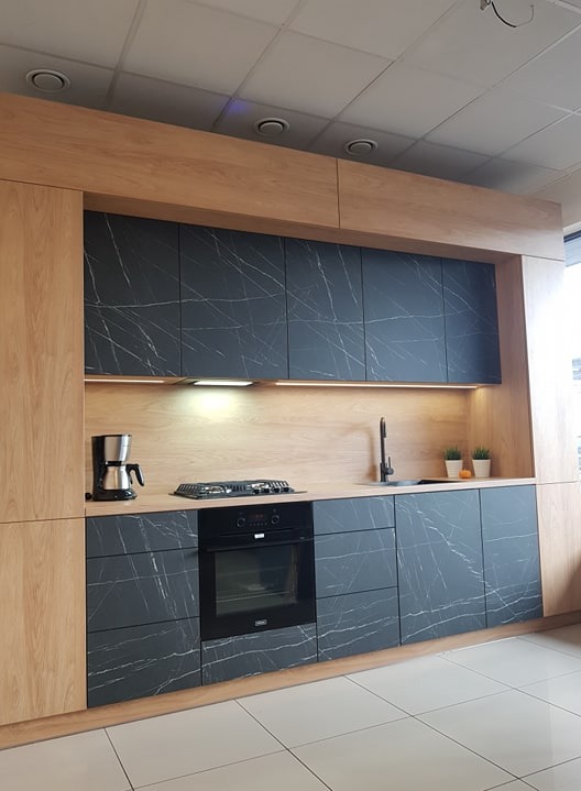 Nowoczesna kuchnia z frontami w kolorze ciemnego marmuru i drewnianymi akcentami, oświetleniem podszafkowym LED, wyposażona w płytę grzewczą, piekarnik i ekspres do kawy.