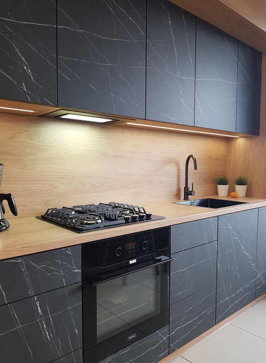 Nowoczesna kuchnia: czarne fronty szafek z marmurkowym wzorem, drewniany blat i panel ścienny, czarna armatura, oświetlenie LED pod szafkami wiszącymi.