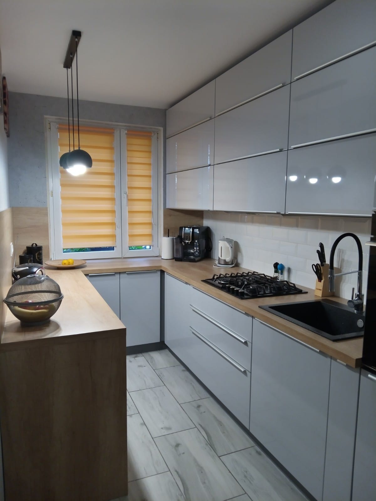 Kompaktowa kuchnia w bloku z jasnoszarymi lakierowanymi frontami i blatem w kolorze dębu Nebraska, z piekarnikiem na wysokości i zmywarką 60 cm, oświetlona lampą wiszącą z trzema kulistymi kloszami.