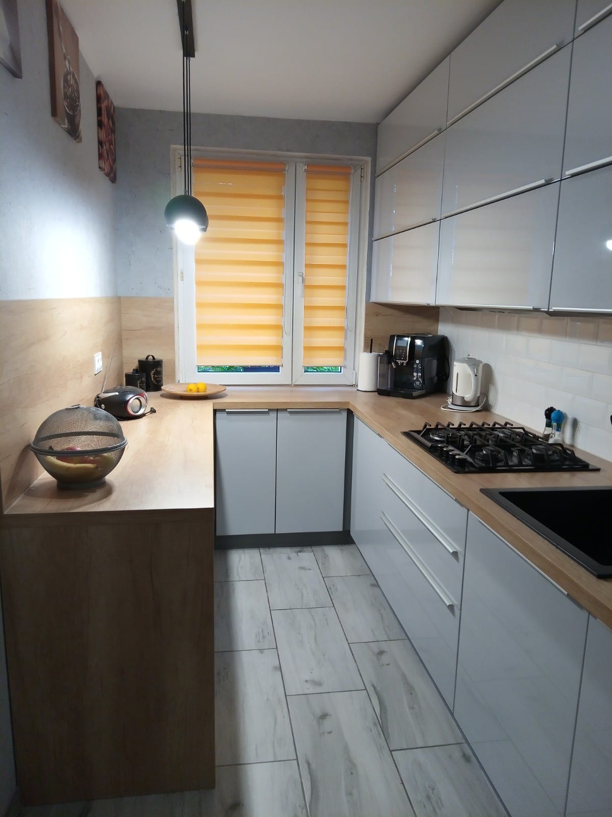 Jasnoszara kuchnia w bloku z lakierowanymi frontami i blatem z dekorem dębu Nebraska, piekarnik na wysokości i zmywarka 60 cm, widok na okno z pomarańczowymi roletami.