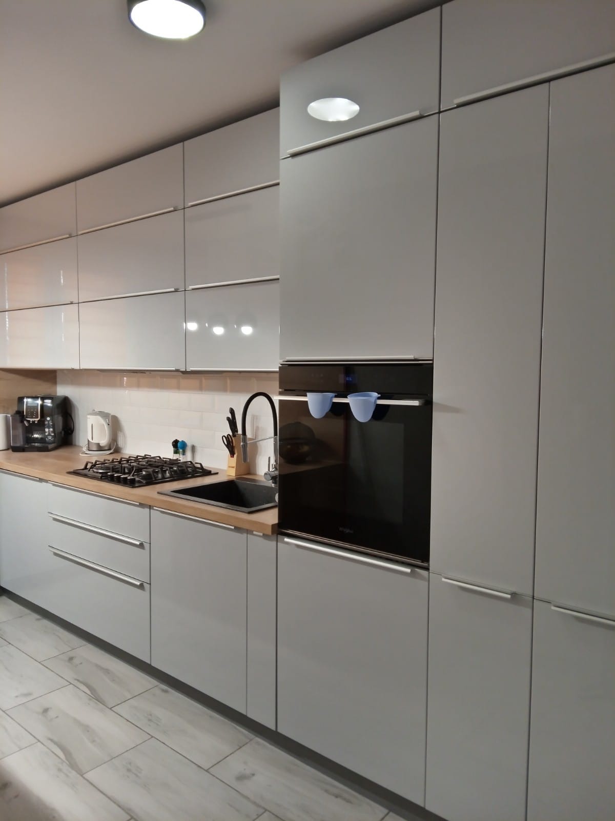 Jasnoszara kuchnia z lakierowanymi frontami i blatem w kolorze dębu Nebraska. Widoczny piekarnik w zabudowie, płyta gazowa i czarny zlew. Na blacie ekspres do kawy i czajnik. Na piekarniku wiszą...