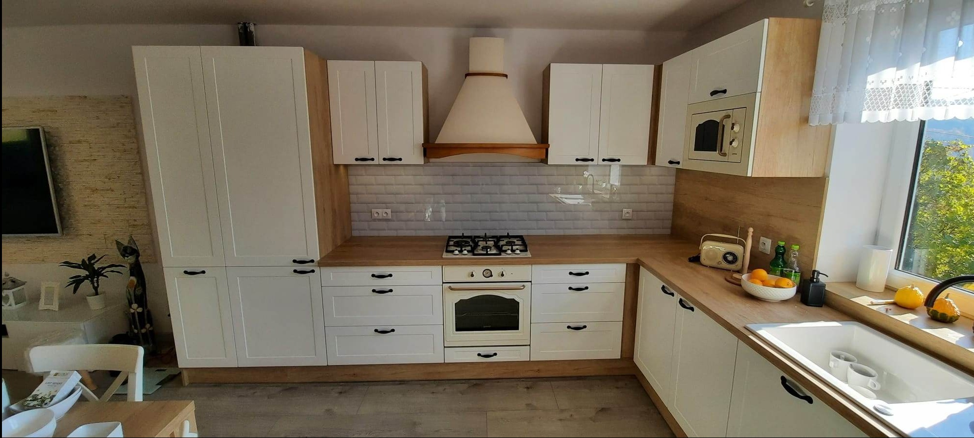 Jasna kuchnia z białymi frontami i blatem w kolorze dębu nebraska, kremowy piekarnik i kuchenka gazowa, styl retro, widok na zewnątrz przez okno.