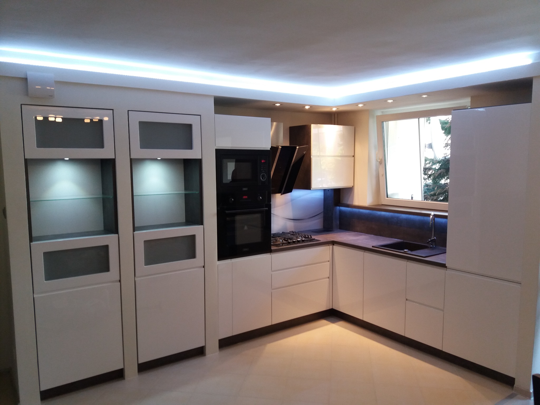 Nowoczesna kuchnia w bieli i szarości, fronty lakierowane na wysoki połysk, frezowane uchwyty, witryny ekspozycyjne z oświetleniem, czarny piekarnik i płyta gazowa, betonowy blat, oświetlenie LED...