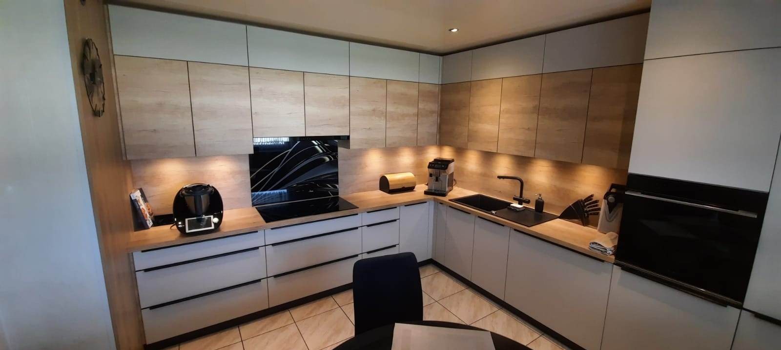 Nowoczesna kuchnia z białymi dolnymi szafkami i górnymi szafkami w kolorze dębu Nebraska, czarne uchwyty, hartowana szyba ze wzorem na ścianie między szafkami, czarny zlew i sprzęt AGD.