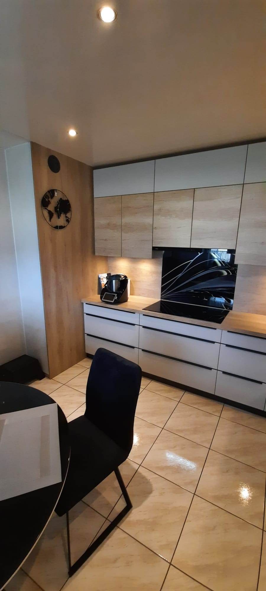 Nowoczesna kuchnia z frontami w odcieniach bieli i drewna, czarnymi uchwytami, płytą indukcyjną i panelem szklanym z abstrakcyjnym wzorem, uzupełniona o zegar ścienny z mapą świata i czarne krzesło...