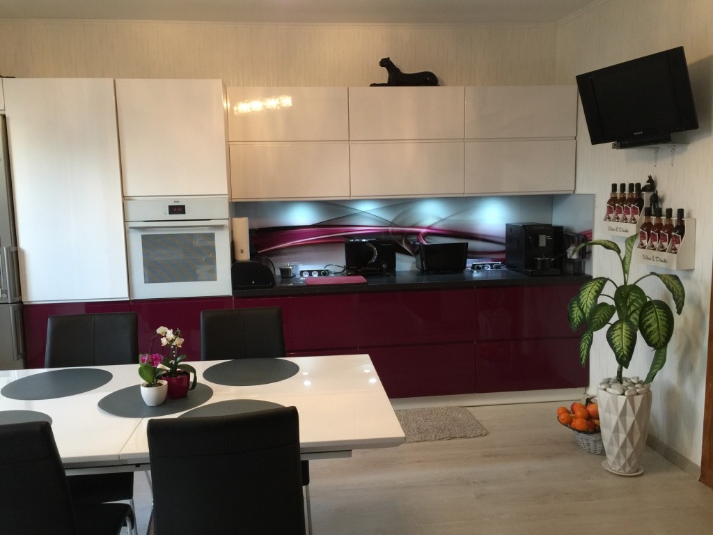 Nowoczesna kuchnia z białymi szafkami górnymi i fuksjowymi szafkami dolnymi na wysoki połysk, czarny blat, szklany panel nad blatem z abstrakcyjnym wzorem, stół z czarnymi krzesłami w centrum...