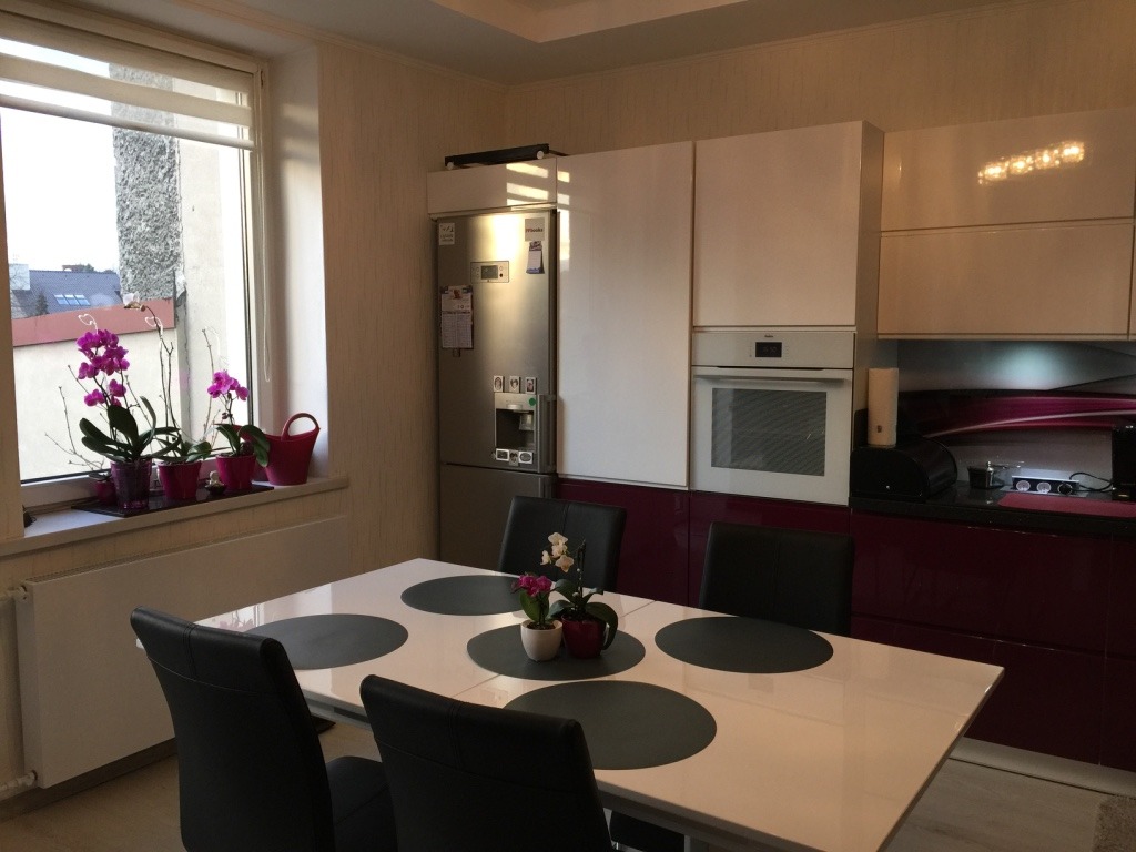 Nowoczesna kuchnia z białymi szafkami górnymi i fuksjowymi dolnymi, frezowane uchwyty, szklany panel na ścianie z subtelnym wzorem, stół z czarnymi podkładkami i krzesłami w tle okno z kwiatami.