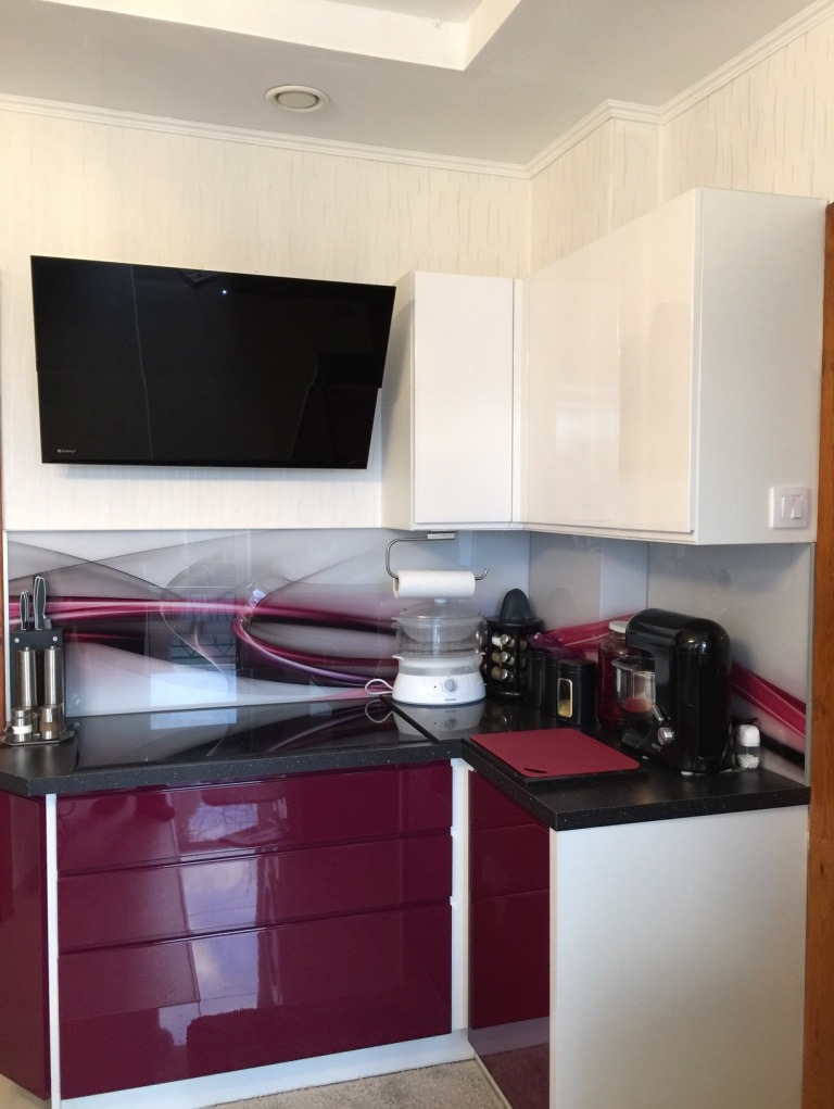 Kuchnia z frontami w kolorze fuksji w wysokim połysku, zestawiona z białymi szafkami, czarnym blatem i szybą hartowaną z abstrakcyjnym wzorem na ścianie.