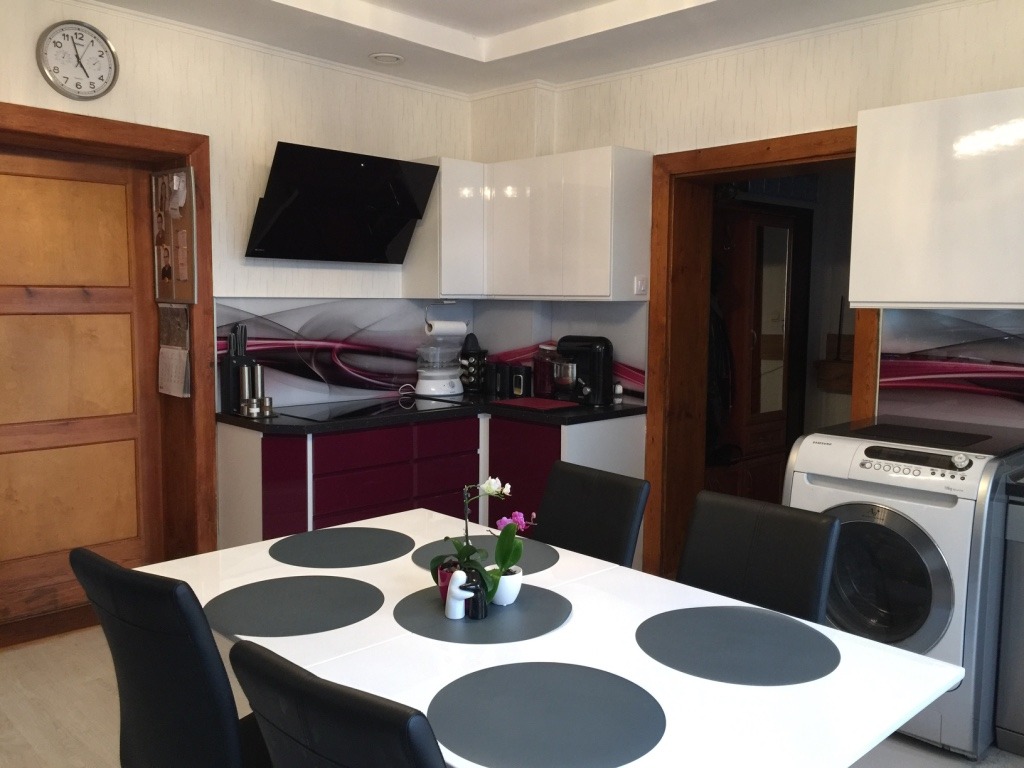 Nowoczesna kuchnia z frontami w kolorze fuksji i białym połysku, frezowanymi uchwytami i szklanym panelem ściennym z abstrakcyjnym wzorem, widok z perspektywy stołu jadalnego.