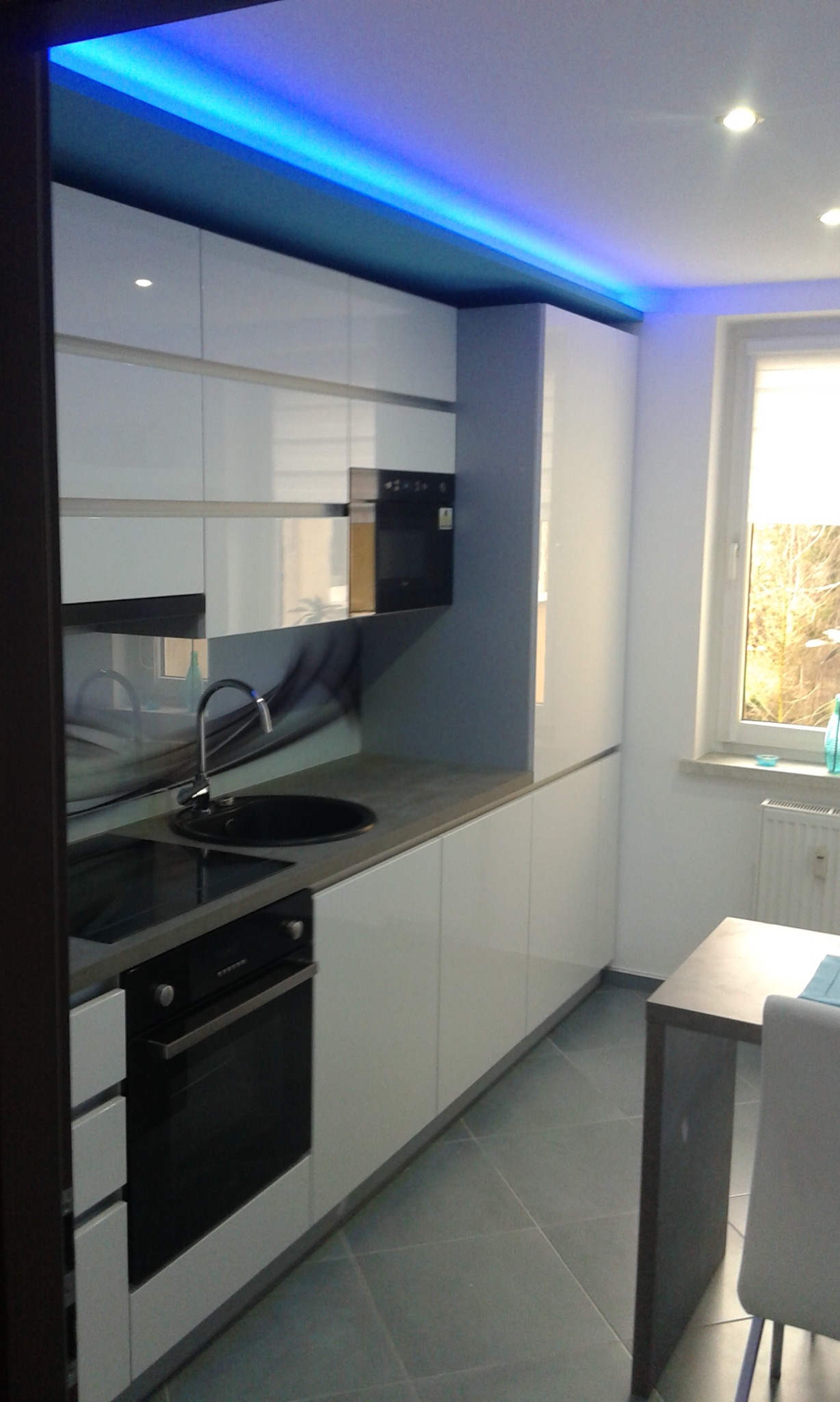 Nowoczesna kuchnia w bloku z białymi lakierowanymi frontami i szarym blatem, oświetlona niebieskim LED-em podszafkowym. Widoczny stół w kolorze blatu i czarny zlew.