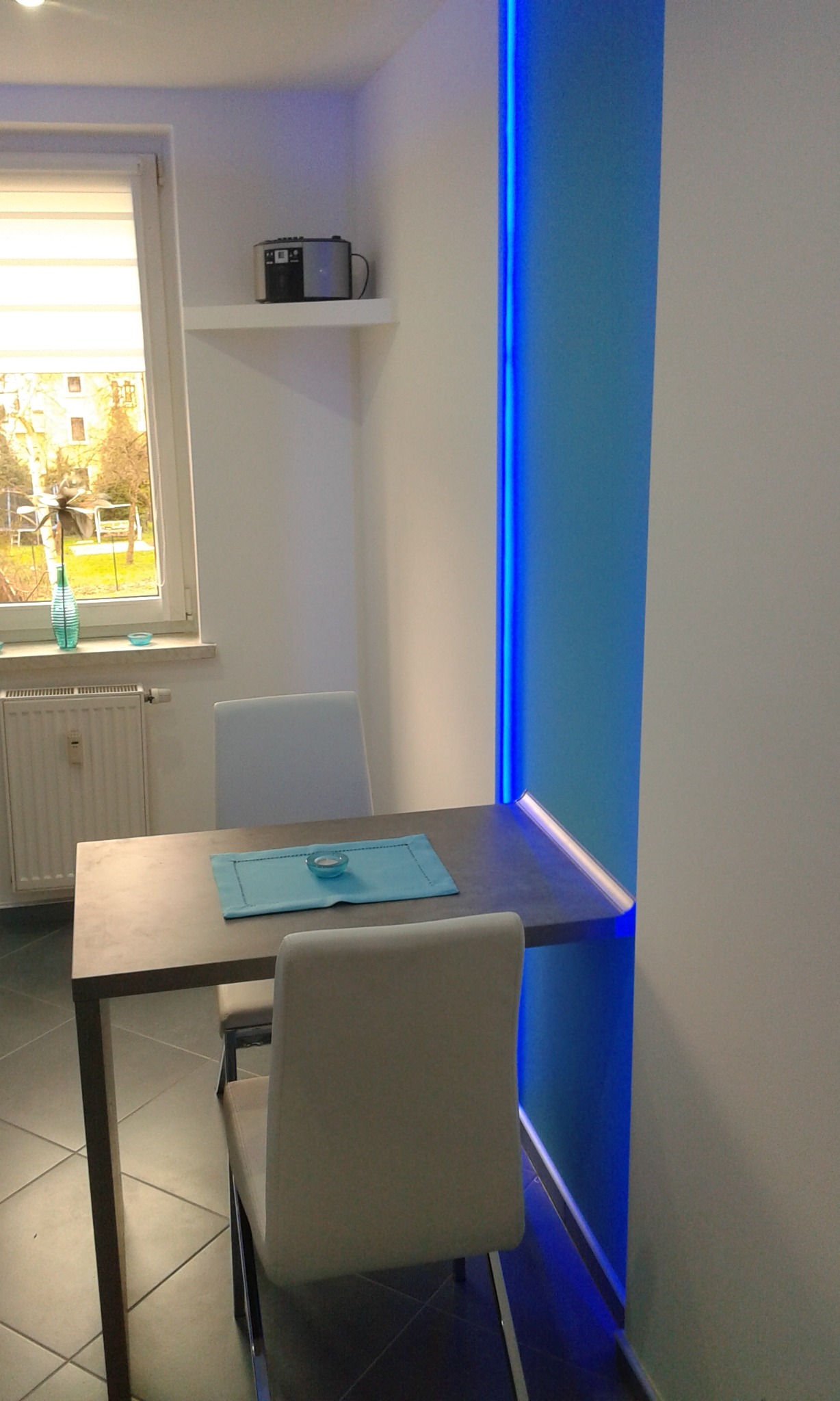 Nowoczesna aranżacja małej kuchni w bloku: Stół z dwoma krzesłami, betonowy blat, niebieskie oświetlenie LED wzdłuż ściany, okno z widokiem na ogród.