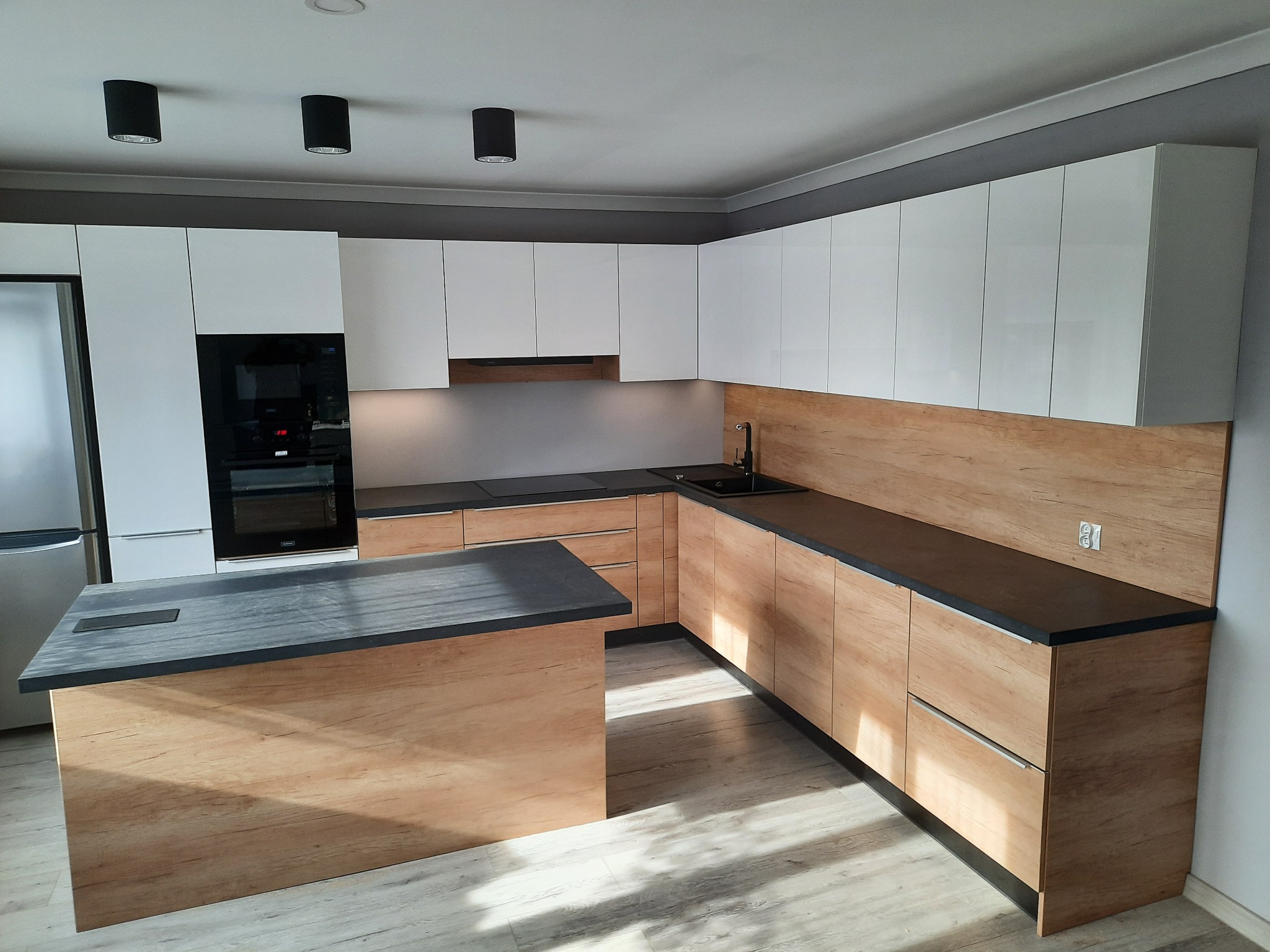 Nowoczesna kuchnia z wyspą: białe górne szafki, dolne szafki i wyspa w odcieniach dębu Nebraska, czarny blat i armatura, oświetlenie punktowe.