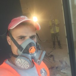 Nikita Bonzhukov - Pracownik w masce ochronnej i kamizelce odblaskowej z czołówką na czapce, w tle drugi pracownik i gruz po remoncie.