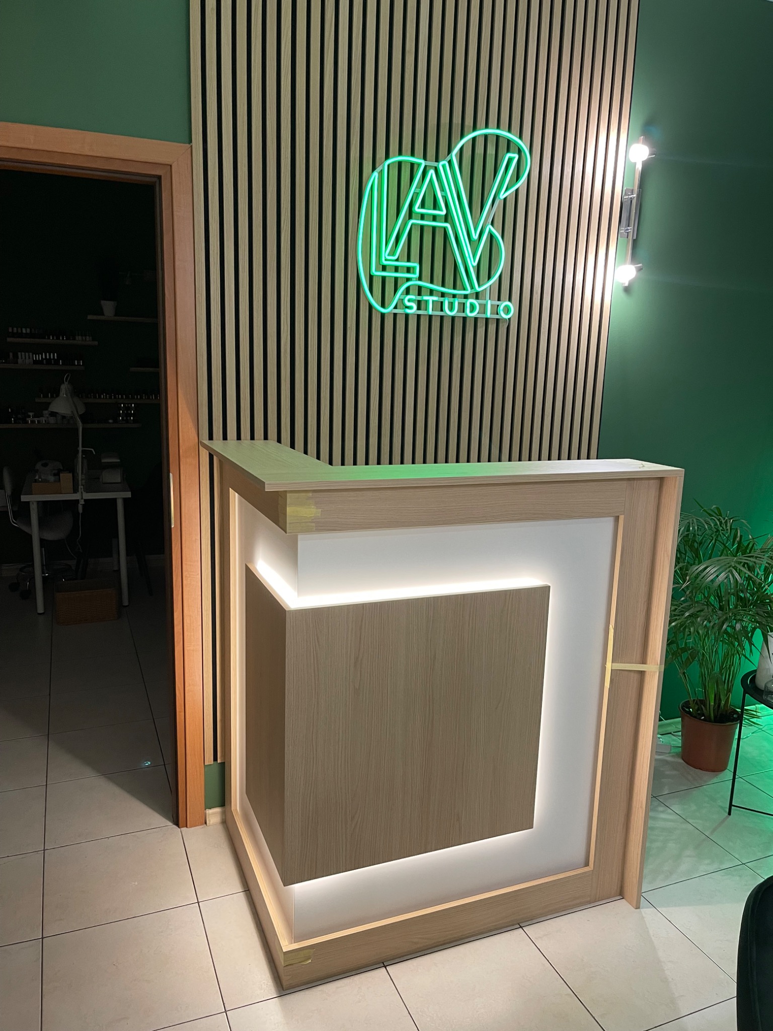 Recepcja w salonie kosmetycznym z podświetleniem LED, zielona ściana i neonowe logo na tle drewnianych paneli ściennych.