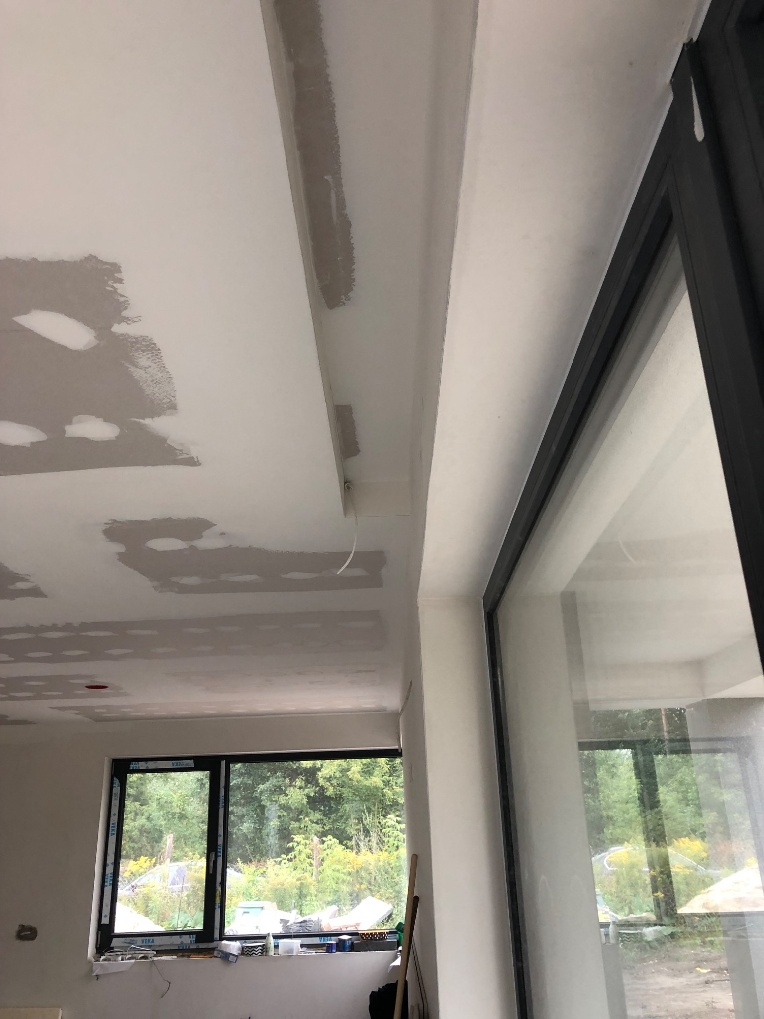 Wnętrze w trakcie prac w Warszawie: sufit po szpachlowaniu, widoczne niedoskonałości i ślady po łatach, okno z widokiem na zieleń.