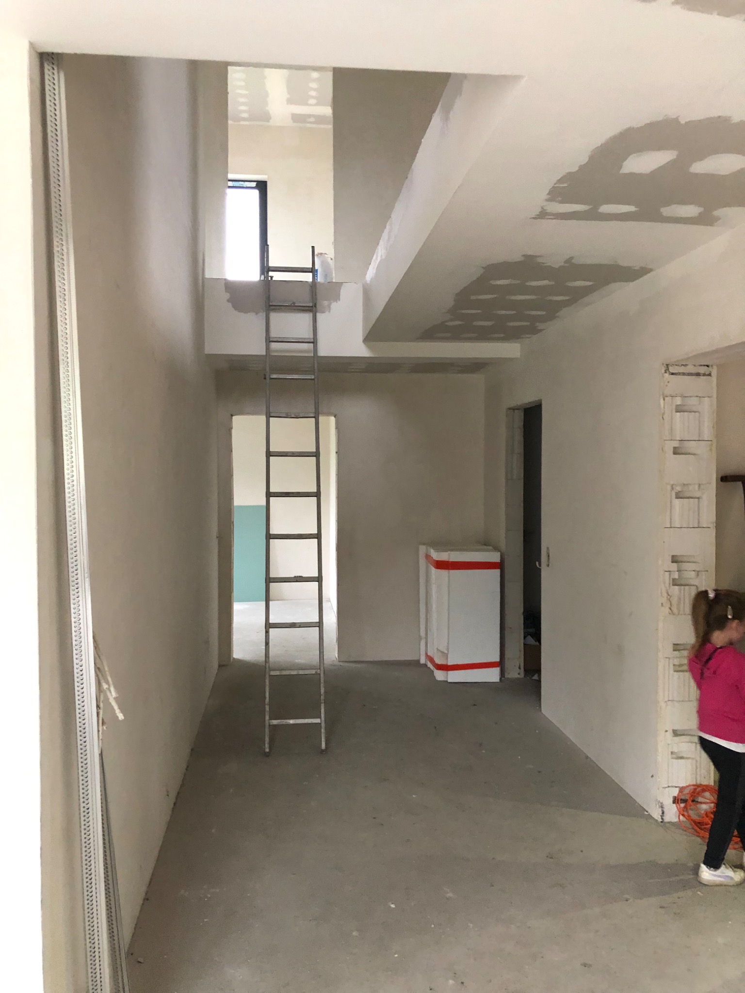 Wnętrze w trakcie prac wykończeniowych: surowe ściany i sufit po szpachlowaniu, aluminiowa drabina oparta o ścianę, widoczny fragment dziecka w prawym dolnym rogu.