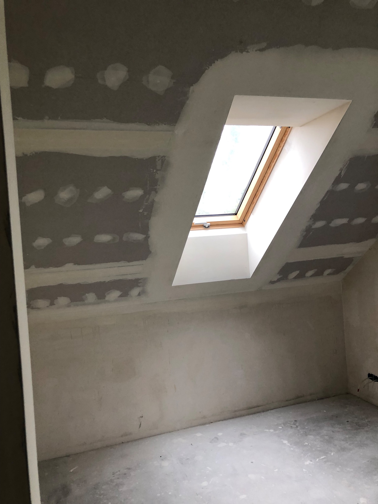 Poddasze w trakcie prac wykończeniowych: ściany i sufit z płyt gipsowo-kartonowych, widoczne miejsca po wkrętach zaszpachlowane, okno dachowe w białej obudowie.