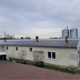 Instalacja paneli słonecznych na dachu budynku gospodarczego z widocznymi silosami zbożowymi w tle, zaparkowany biały bus z otwartymi drzwiami sugeruje trwające prace.