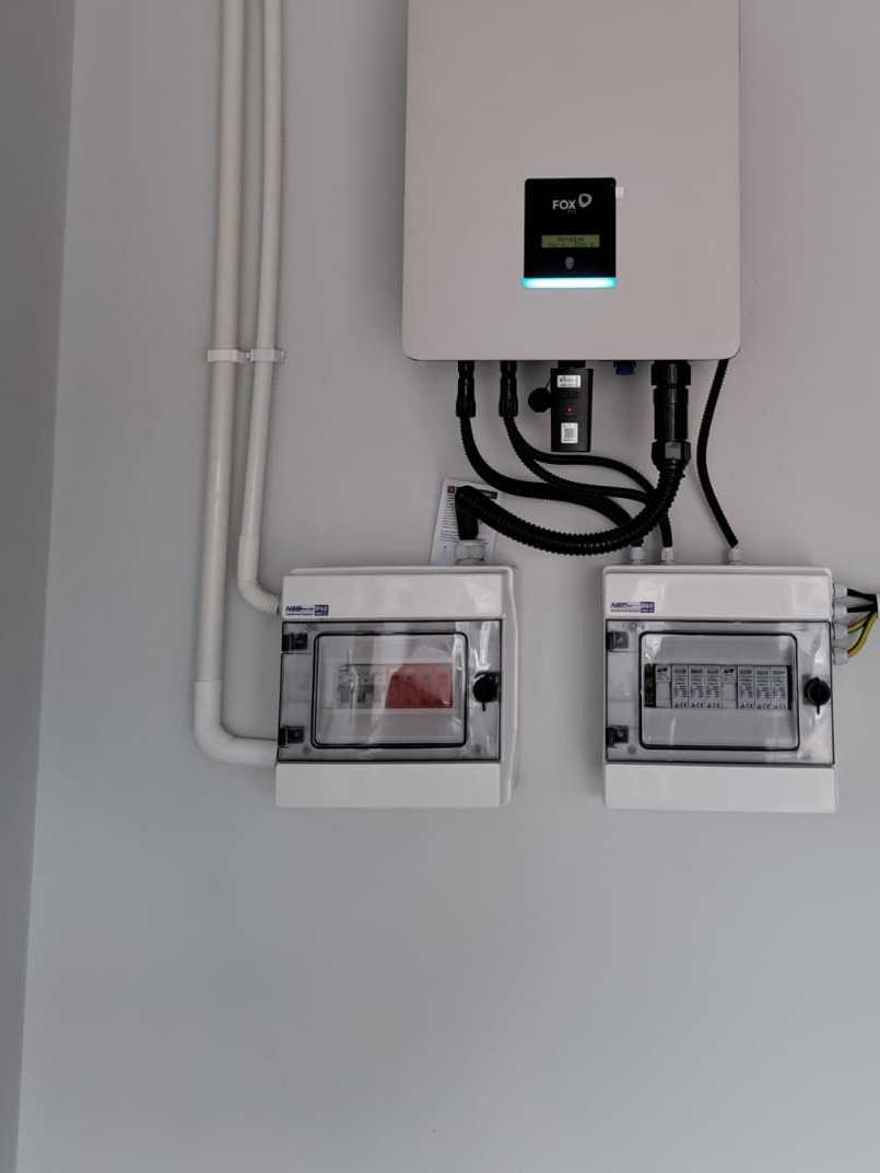 Biały inwerter marki FOX z podłączonymi kablami i dwoma rozdzielnicami na jasnej ścianie.