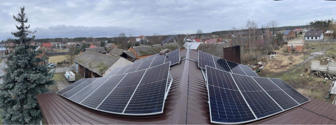 Instalacja fotowoltaiczna na dachu domu jednorodzinnego w wiejskiej scenerii, widok z góry na panele słoneczne i okoliczne budynki, pochmurne niebo.