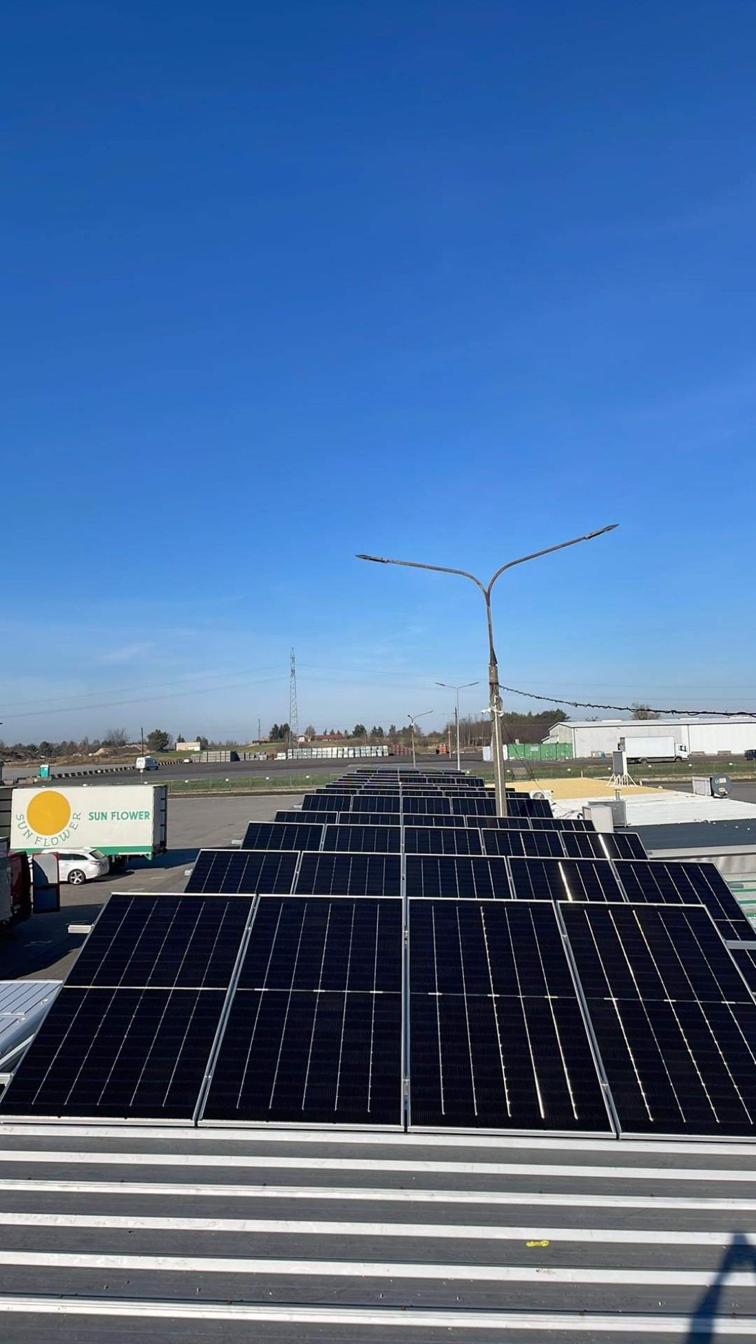 Rząd paneli słonecznych na dachu budynku, w tle błękitne niebo, słup oświetleniowy i zaparkowana ciężarówka z logiem Sun Flower.