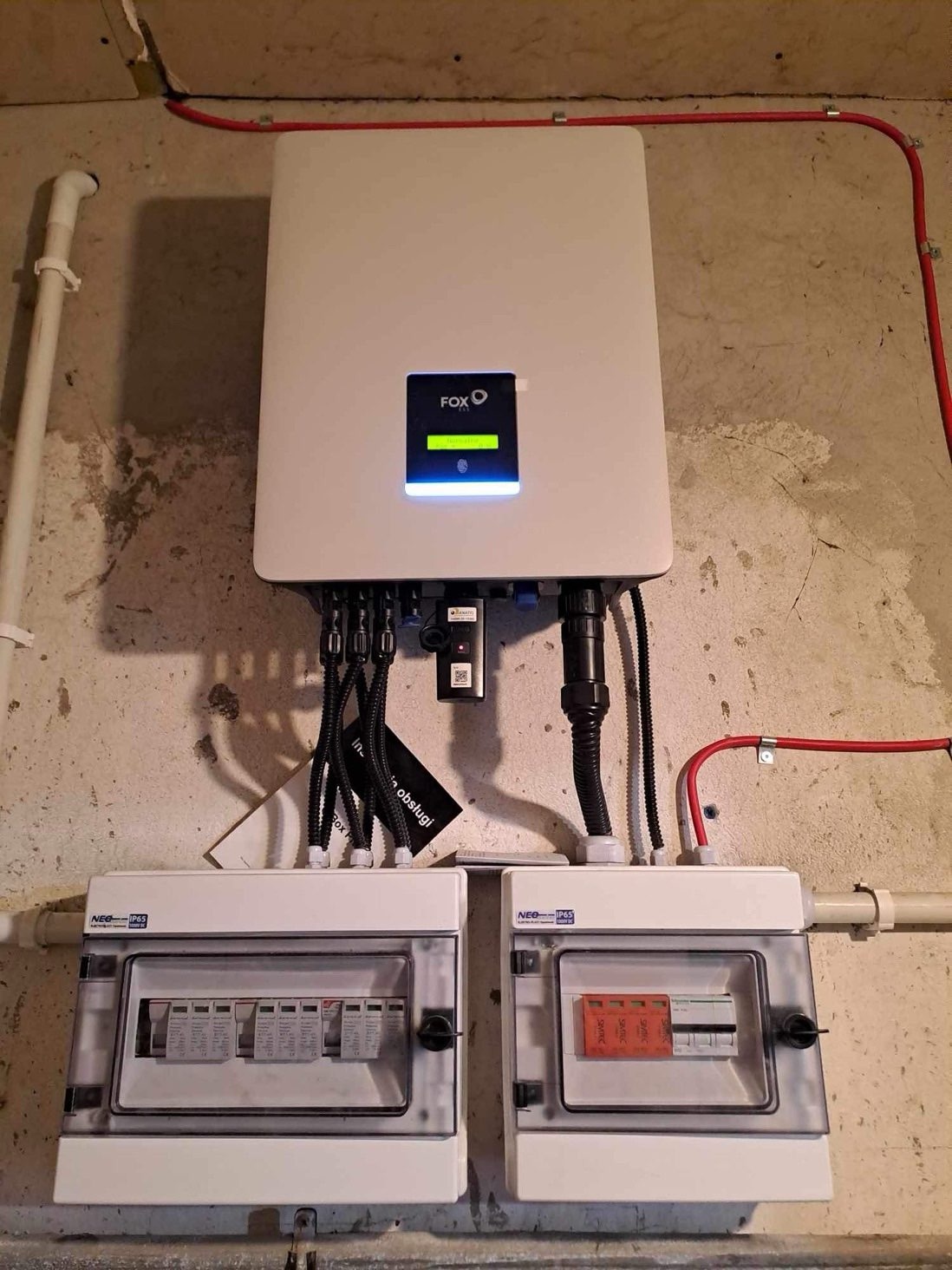 Biały inwerter marki FOX z podłączonymi kablami, zamontowany na ścianie nad dwiema skrzynkami z bezpiecznikami, widoczne czerwone kable elektryczne prowadzone po ścianie.