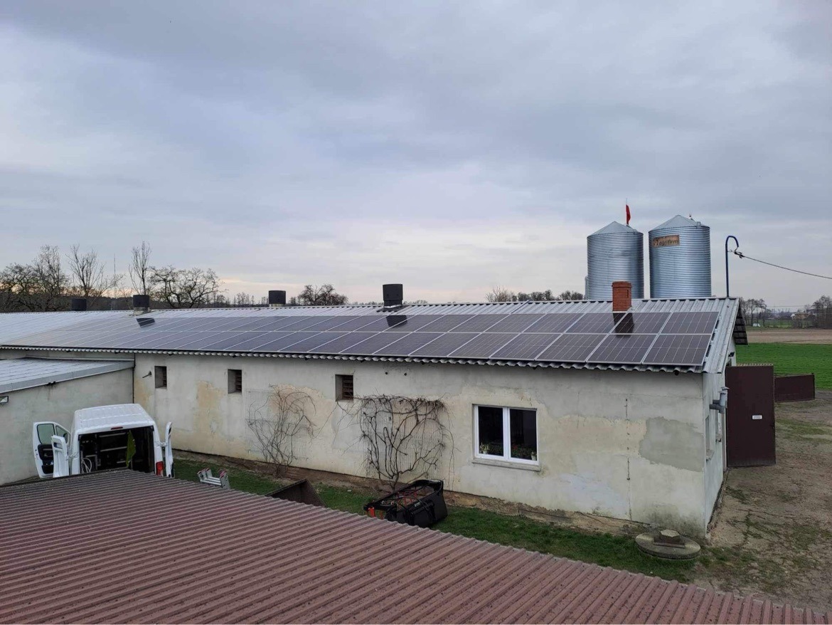 Instalacja paneli słonecznych na dachu budynku gospodarczego z widocznymi silosami zbożowymi w tle, zaparkowany biały bus z otwartymi drzwiami sugeruje trwające prace.