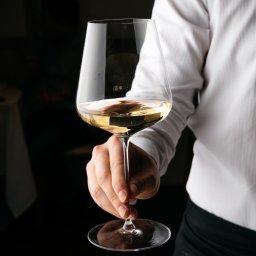 Ilia Aleszkowski - Kelner w białej koszuli trzyma kieliszek białego wina na tle ciemnego wnętrza restauracji.