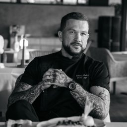 Ilia Aleszkowski - Czarno-białe portretowe zdjęcie mężczyzny z brodą i tatuażami, ubranego w czarną koszulę z napisem 'Dmitriy Dulov Brand Chef', siedzącego przy stole z talerzem jedzenia w restauracji.