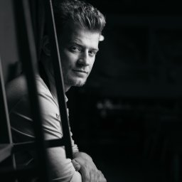 Ilia Aleszkowski - Portret mężczyzny w czerni i bieli, lekko zamyślony, z dłońmi splecionymi na oparciu krzesła, widziany przez metalową konstrukcję, z ciemnym tłem.