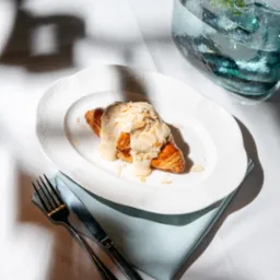 Kremowy rogalik z płatkami migdałów na białym talerzu, obok sztućce i dzbanek z wodą, na tle białego obrusu i jasnoniebieskiej serwetki.