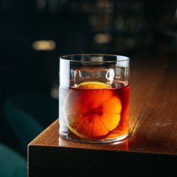 Ilia Aleszkowski - Kieliszek z bursztynowym płynem, kostką lodu i plasterkiem pomarańczy na drewnianym blacie baru, w tle rozmyte światła.