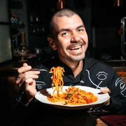 Ilia Aleszkowski - Uśmiechnięty mężczyzna z kolczykiem w nosie, w czarnej koszuli z białymi wzorami, trzyma widelec z makaronem spaghetti nad talerzem pełnym spaghetti z sosem pomidorowym w restauracji.