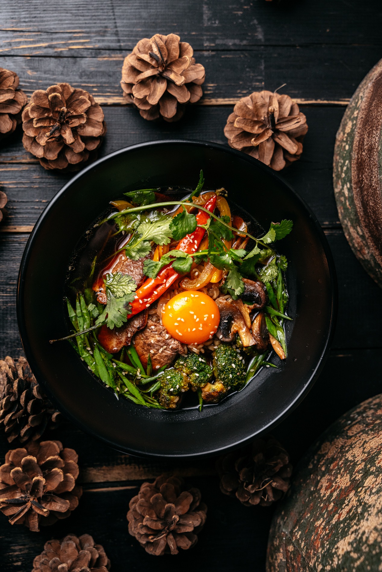Czarna miska z azjatyckim daniem: wołowina, warzywa (brokuły, papryka, pieczarki), zielony szczypior, kolendra i żółtko, posypane sezamem, na ciemnym drewnianym tle z szyszkami i dynią.