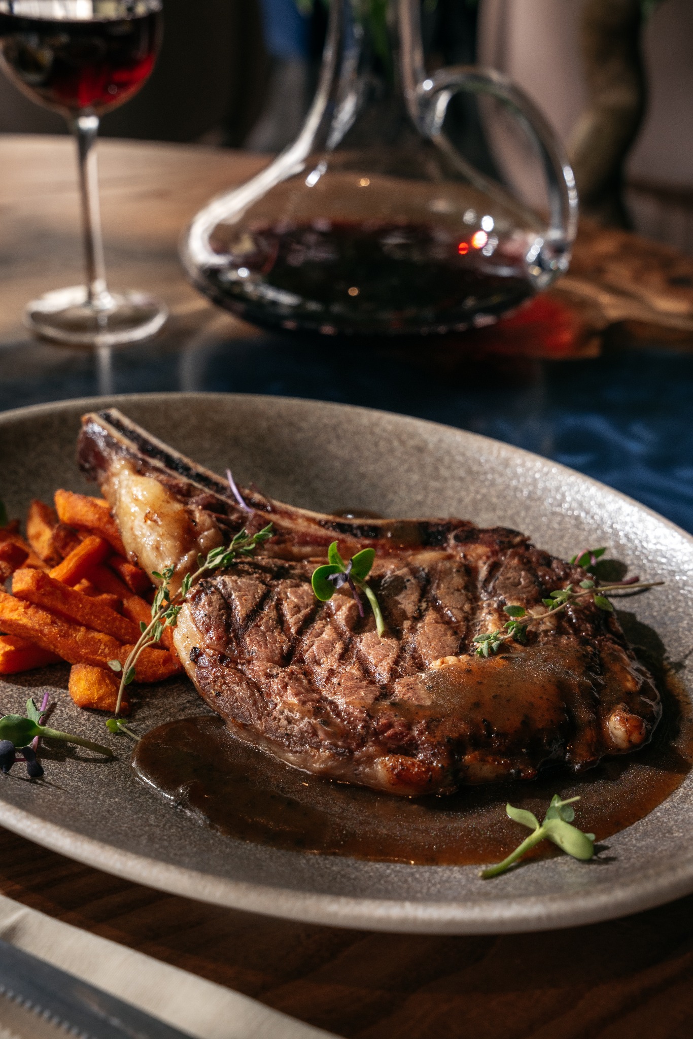 Grillowany stek z kością podany na szarym talerzu z frytkami z batatów, dekorowany świeżymi ziołami, w tle karafka i kieliszek czerwonego wina.
