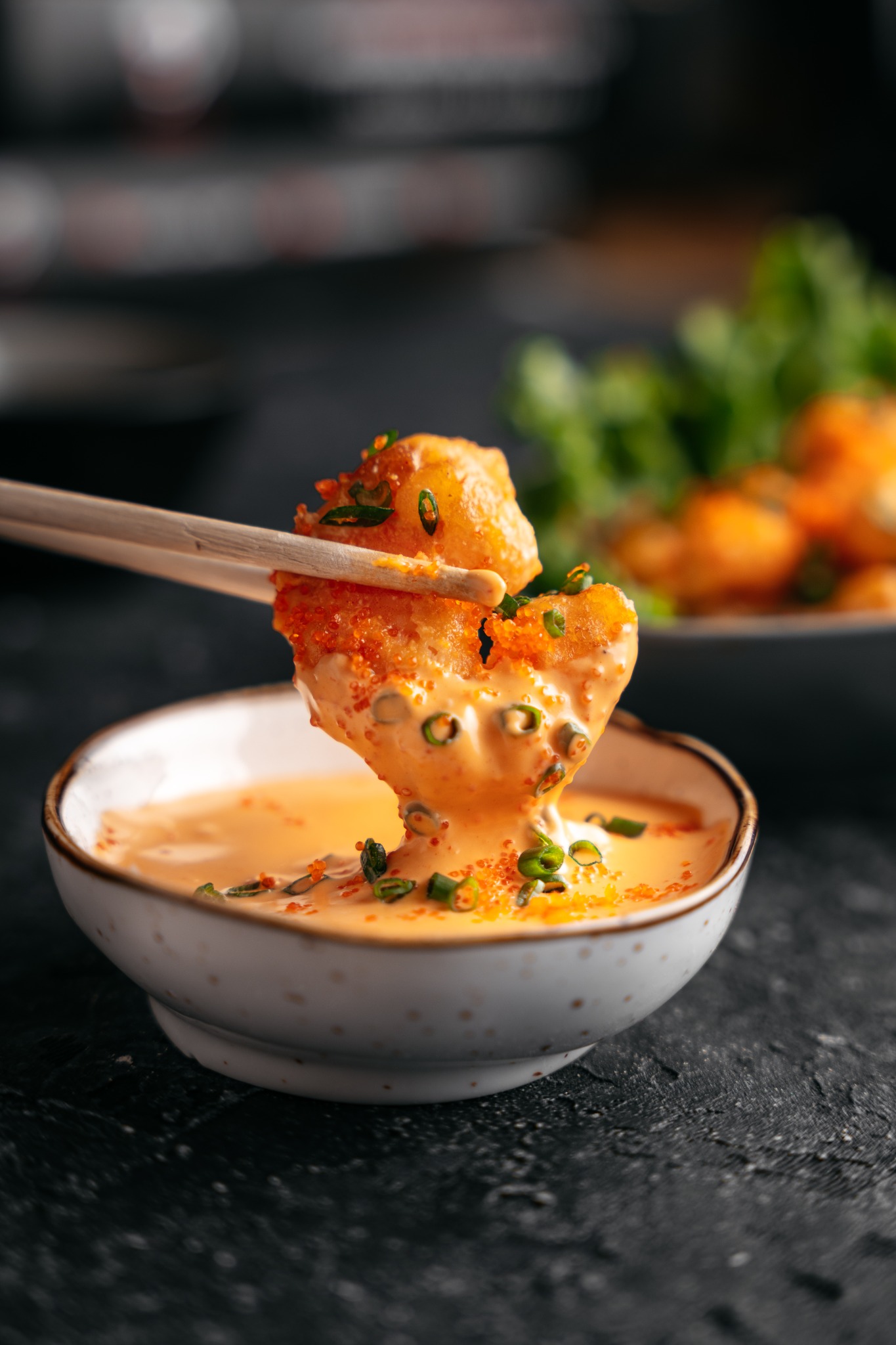 Krewetka w tempurze trzymana pałeczkami zanurzana w kremowym sosie z posypką z zielonego szczypiorku i pomarańczowego kawioru, na tle miseczki z sosem i talerza z zieloną sałatą.