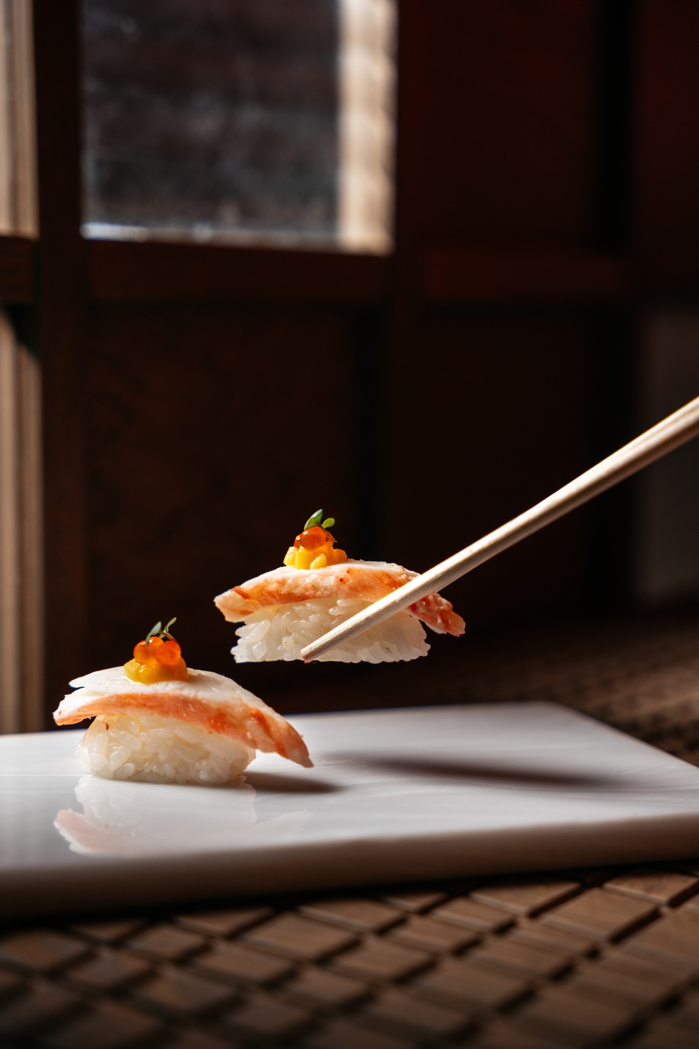 Dwa kawałki sushi nigiri z krabem, ikrą i mikro-ziołami, jeden podnoszony pałeczkami, na białym talerzu z rozmytym tłem okna i bambusowej maty.