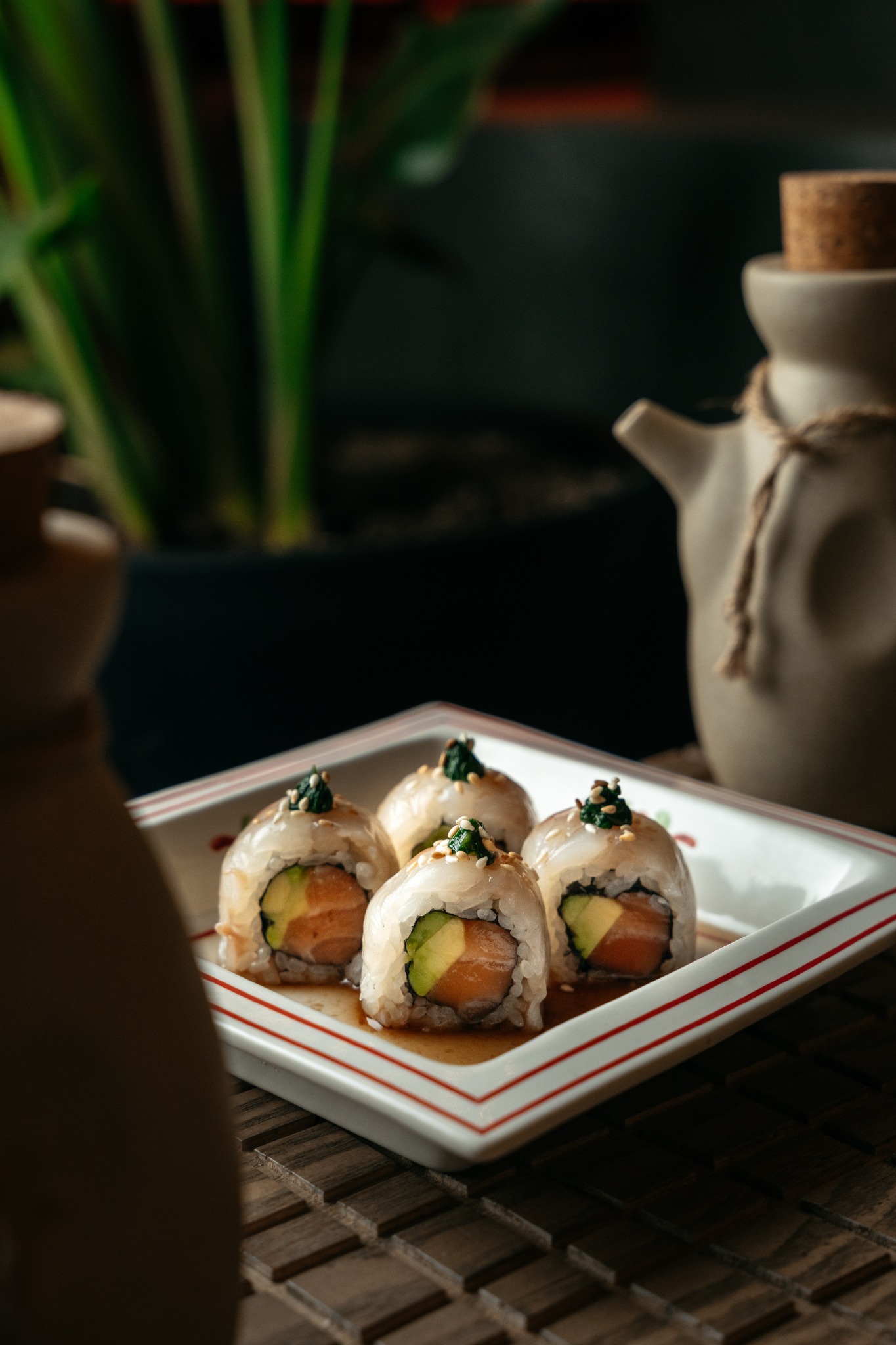 Cztery rolki sushi z łososiem i awokado, polane sosem i posypane sezamem i zieloną dekoracją, ułożone na białym talerzu z czerwoną obwódką, w tle dzbanek i roślina doniczkowa.
