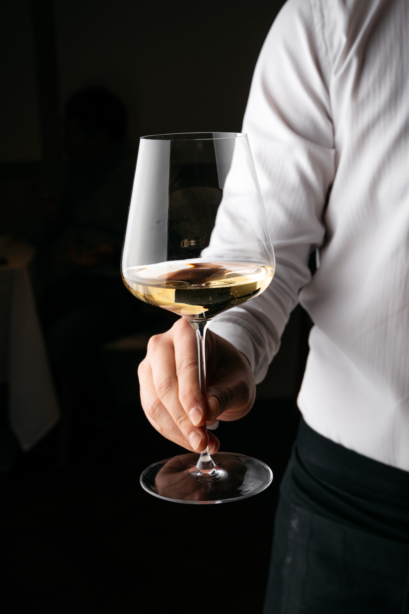 Kelner w białej koszuli trzyma kieliszek białego wina na tle ciemnego wnętrza restauracji.
