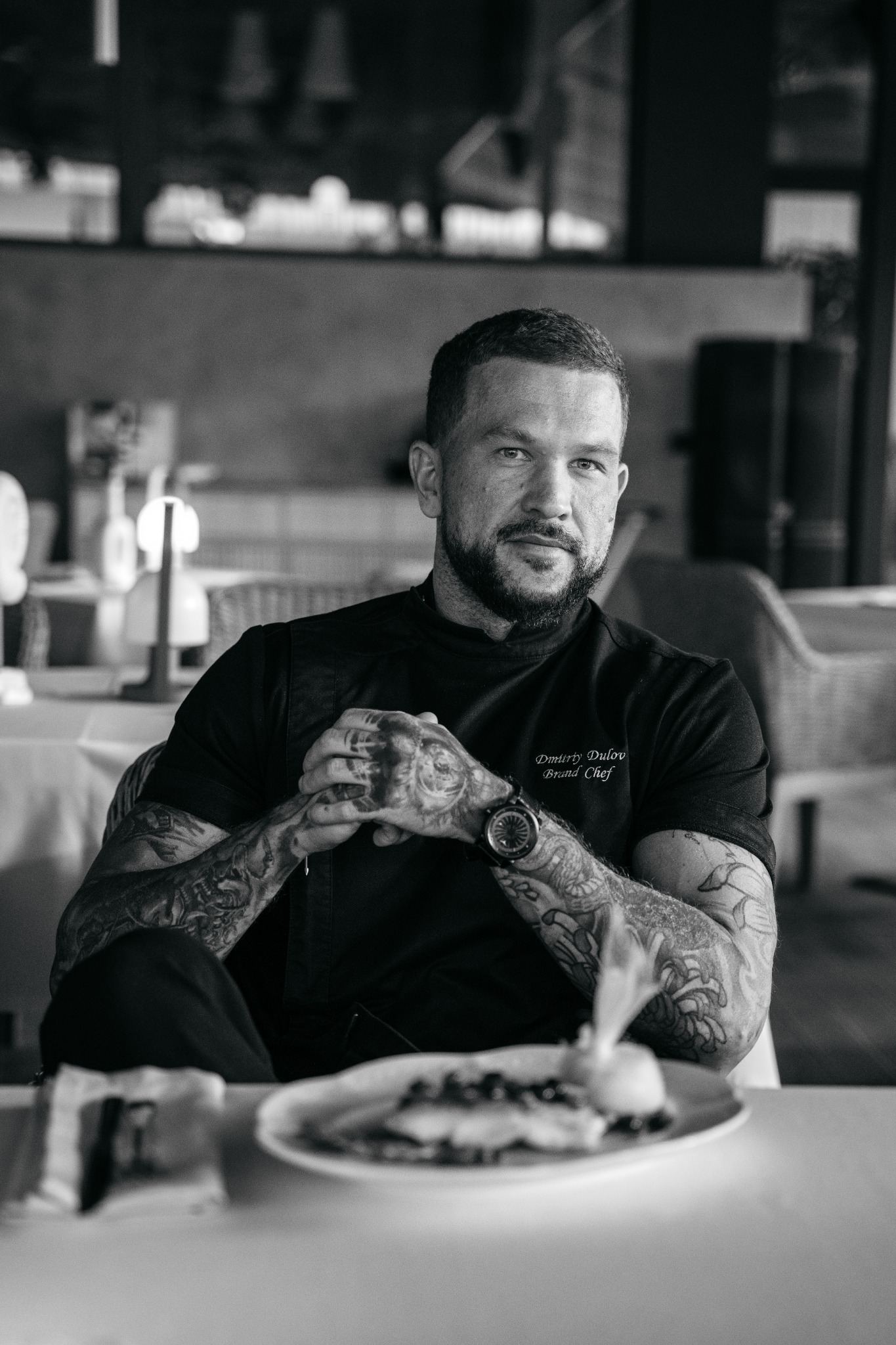Czarno-białe portretowe zdjęcie mężczyzny z brodą i tatuażami, ubranego w czarną koszulę z napisem 'Dmitriy Dulov Brand Chef', siedzącego przy stole z talerzem jedzenia w restauracji.
